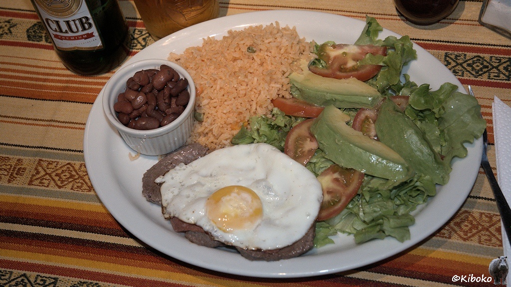 Das Bild zeigt einen Teller mit Steak, auf dem ein Spiegelei liegt. Dazu gibt es reis, Salat, Tomaten, Avocado und ein schälchen Bohnen. Von der Bierflasche ist nur der untere Teil mit der Aufschrift Club sichtbar.
