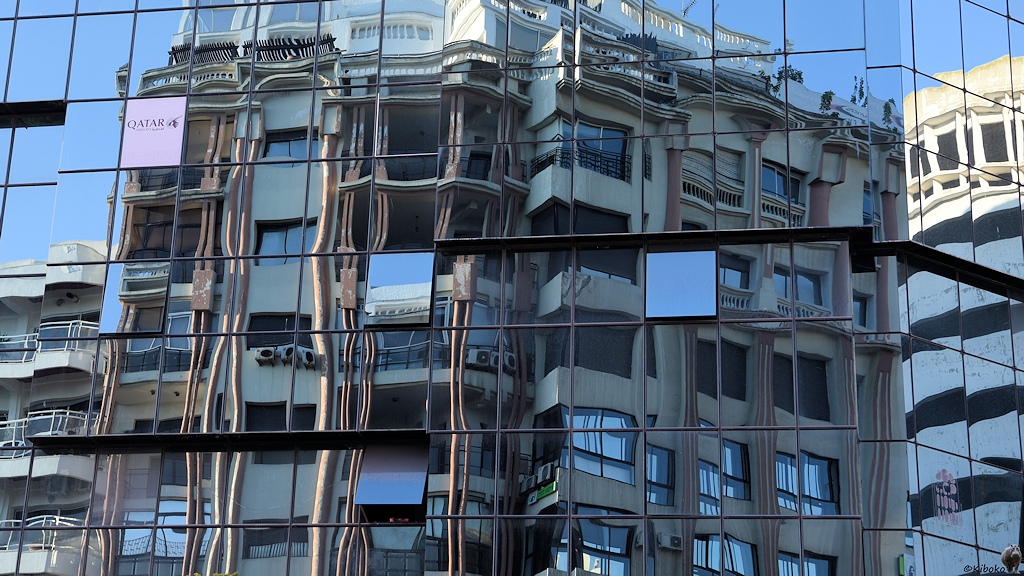 Das Bild eine Glasfassade eines Hauses. Darin spiegelt sich ein mehrgeschossiges Stadthaus. Die Glasscheiben sind nicht ganz plan. Dadurch wird die Spiegelung verzerrt. Das Spiegebild ist unterbrochen durch leicht gekippte Fenster.