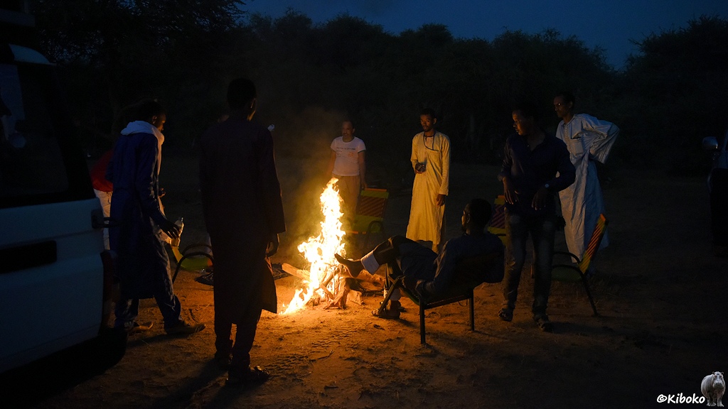 Das Bild zeigt sieben Männer um ein Lagerfeuer. Ein Mann sitzt auf einen Klappstuhl. Die anderen stehen um das Feuer. Das Feuer odert hoch. Ein Mann am linken Bildrand hält eine Plastikflasche mit einer brennbaren Flüssigkeit in der Hand.