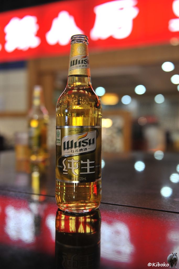 Das Bild zeigt eine druchsichtige, volle Bierflasche mit einem Goldenen Etikett. Darauf steht neben chinesischen Schriftzeichen: Wusu. Der Hintergrund ist unscharf mit roten Bannern und bläulichen Lichtpunkten.