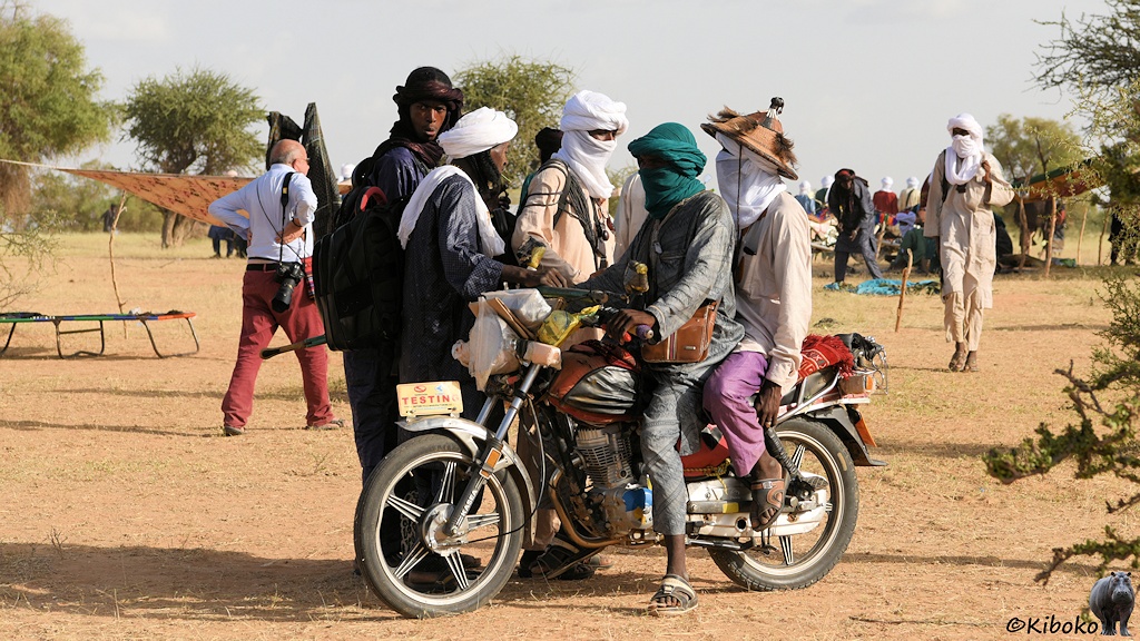 Das Bild zeigt zwei vermummte Männer auf einem Motorrad. Daneben stehen 3 weitere Männer mit Turban. Im Hintergrund sind offene Zelte, eine Liege und vereinzelte kleine Bäume