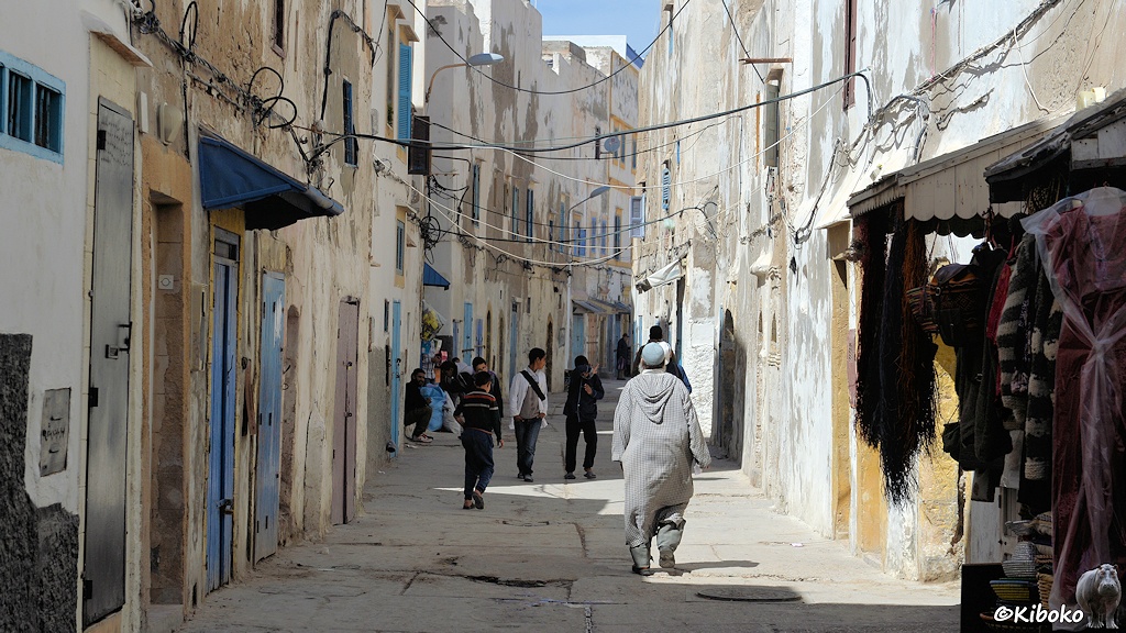 Das Bild zeigt eine Gasse mit unebenenBoden zwischen heruntergekommenen weißen Häusern mit blauen Türen und Fenstern. Auf der Gasse laufen ein Mann im grauen Mantel und jugendliche. Elektroleitungen hängen über der Gasse. Am rechten Bildrand hängen Mäntel als Auslage von einem Geschäft.