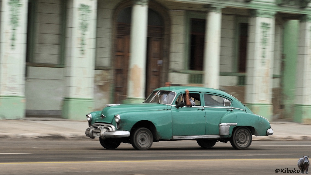 Das Bild zeigt einen Mitzieheraufnahme von einem alten grünen amerikanisches Auto. Das Auto hat ein Fliessheck und einen großen rundlichen hinteren Radkasten. Im Hintergrund ist ein unscharfes, grünes Haus mit Säulen.