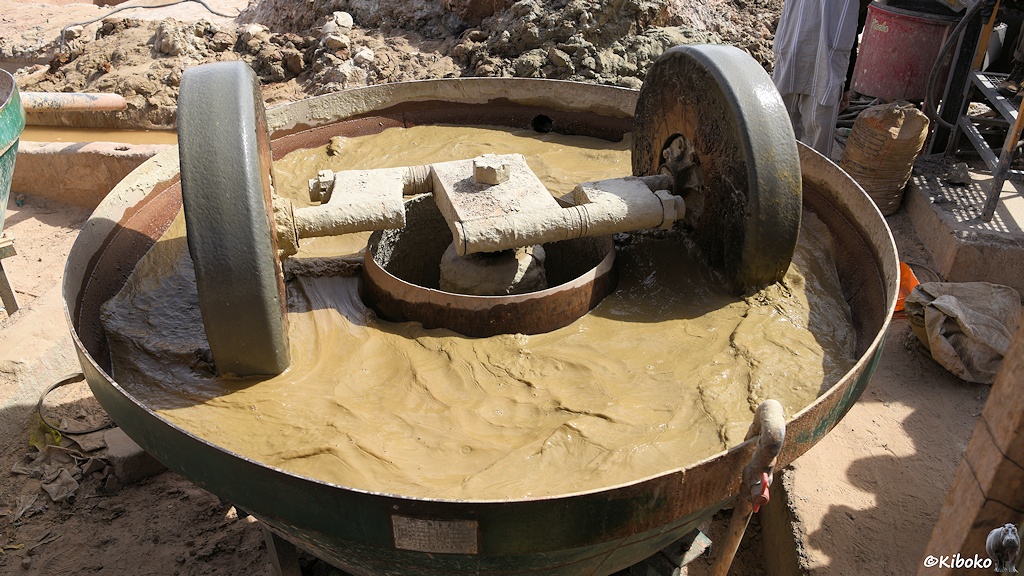 Das Bild zeigt einen Bottich von oben. Darin laufen zwei große Steinräder mit ca. 1m Durchmesser und 20cm Breite im Kreis. Im Bottich schwapt grünlich-bräunliches Wasser.