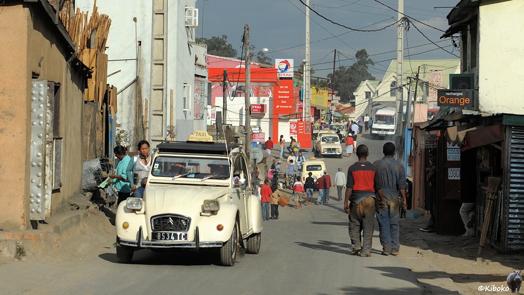 Ein beigefarbener Citroen 2CV kommt als Taxi entgegen. Im Hintergrund stehen mehrere Taxis vom Type Renault R4