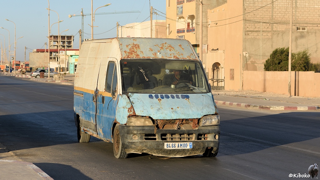 Das Bild zeigt einen verbeulten, verrosteten Kleintransporter ohne Rückspiegel, ohne Kühlergrill auf einer Teerstraße. Die Front ist hellblau. Die Seiten sind oben weiß und unten dunkelblau mit gelber Trennlinie.