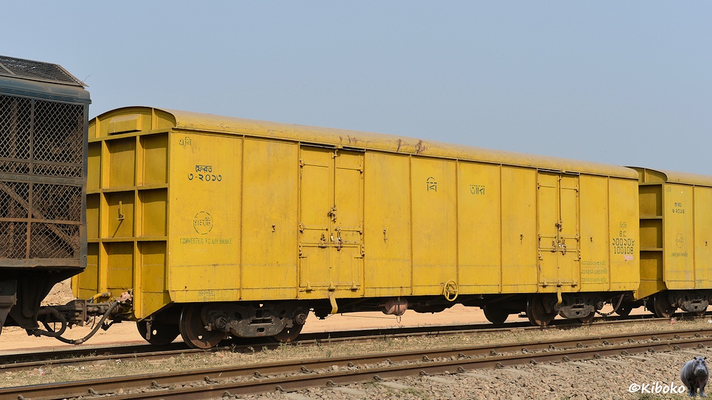 Das Bild zeigt einen gleben vierachsigen gedeckten Güterwagen. Es ist der erste Wagen im Zug. Links sind die Lüfter einer blaugrünen Diesellok sichtbar. Rechts steht ein weiterer Wagen des selben Types.
