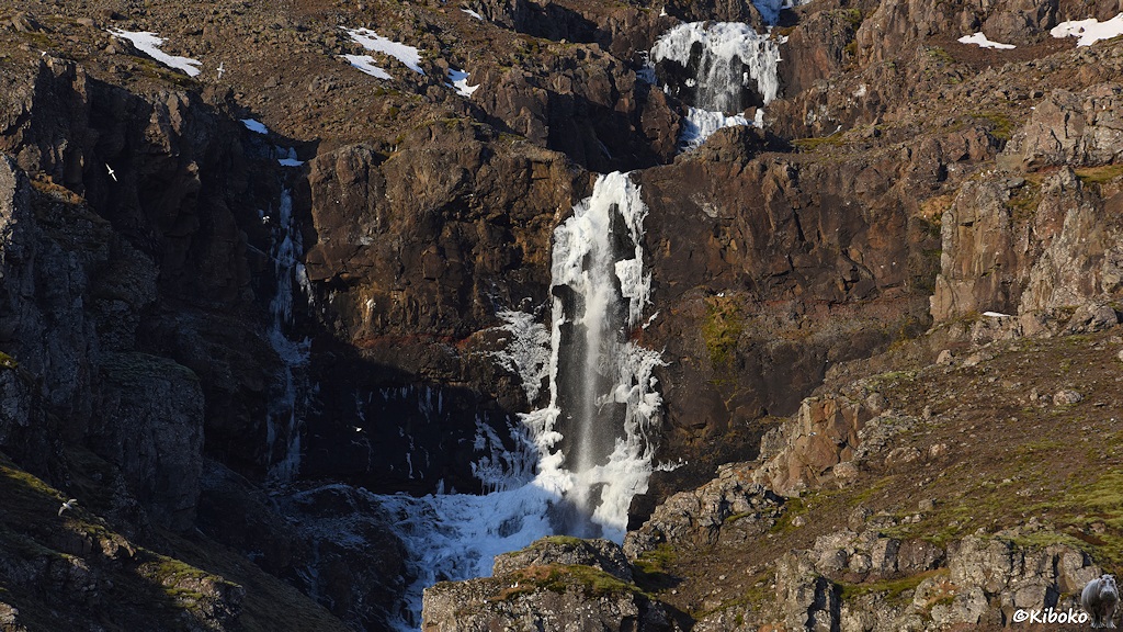 Ein Bach stürzt in zwei Kaskaden über graubraunen Fels. Die Seiten des Wasserfalls sind vereist und bilden Eiszapfen. Nur noch in der Mitte fällt noch Wasser.
