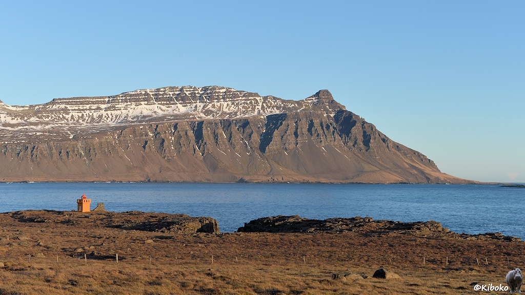 Ein kleiner, viereckiger, oranger Leuchtturm steht an der Einfahrt eines Fjordes mit blauem Wasser. Am gegenüberliegenden Ufer ist eine mächtige Felswand