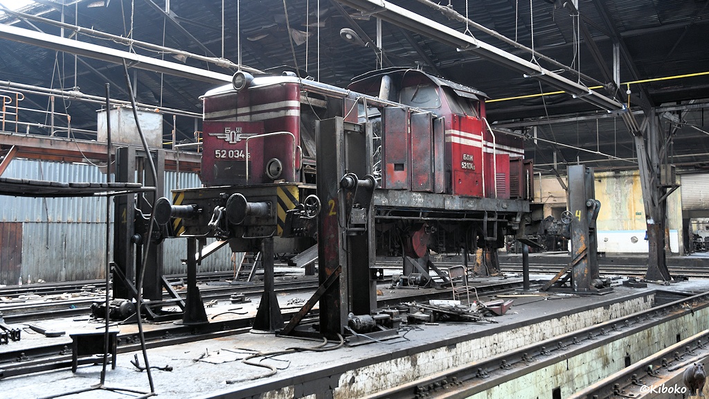 Das Bild zeigt eine rote Diesellokomotive mit Mittelführerstand in einer Werhalle. Die Lok ist aufgebockt und hat keine Radsätze. Alle Türen und Klappen an den Vorbauten stehen offen.