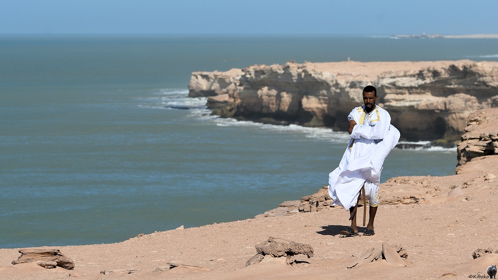 Das Bild zeigt einen Mann im weißen Gewand an einer Abbruchkante am Meer. Die Abbruchkante aus weißem Kalkstein umschließt eine Bucht. Die Wellen treffen auf die senkrechte Felswand.