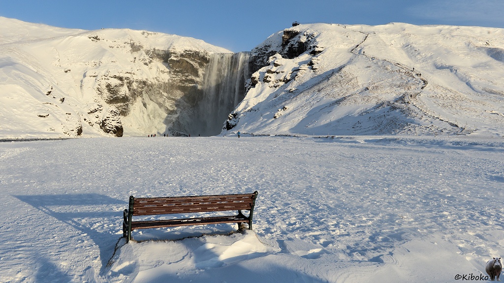Das Bild zeigt eine braune Parkbank von hinten. Dahinter ist ein großer verschneiter Platz. Im Hintergrund ist eine Hügelkette. In der Bildmitte stürzt ein breiter Wasserfall die Bergkette herunter.