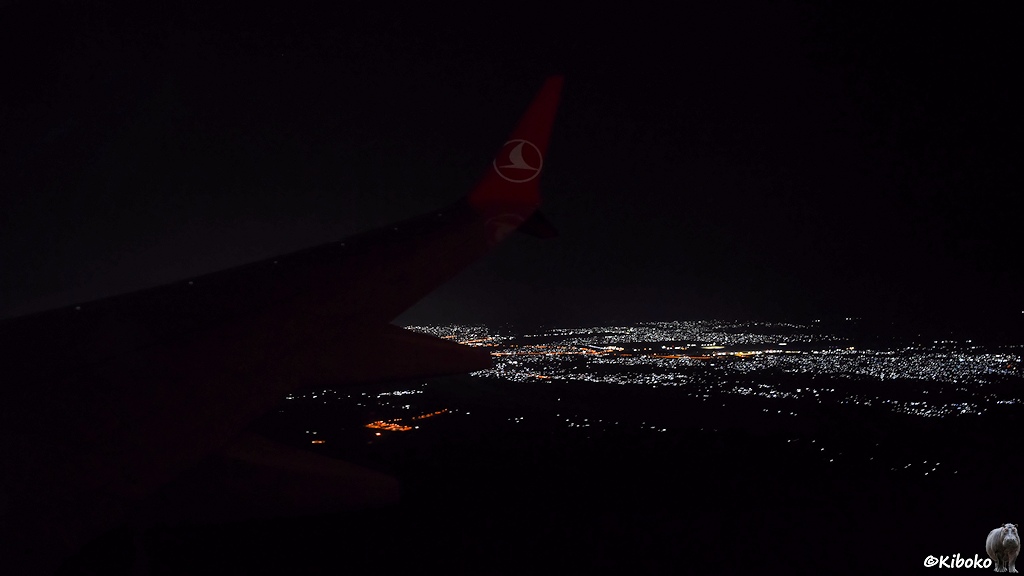 Das Bild zeigt einen nächtlichen Blick aus den Flugzeugfenster auf die Lichter einer Stadt. Schwach ist die rechte Tragfläche sichtbar.
