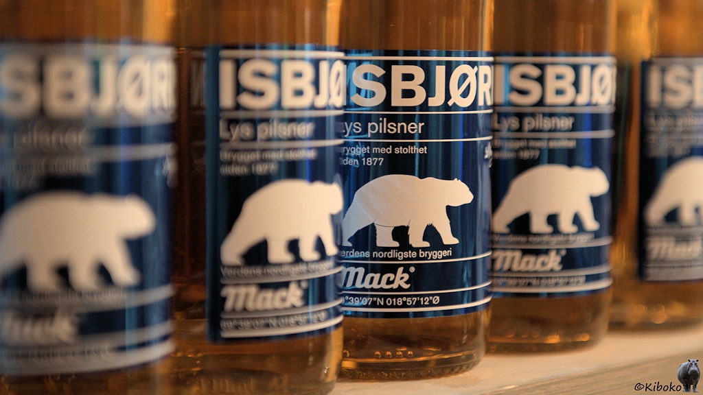 Das Bild zeigt fünf klare Bierflaschen mit ihren Etiketten. Nur die mittlere Flasche ist scharf abgebildet. Das Etikett zeigt einen weißen Eisbär im Profil auf dunkelblauem Hintergrund. Mit weißer Schrift stehen unter anderem die Biersorte Isbjørg, der Brauereiname Mack und die Koordinaten auf dem Etikett.