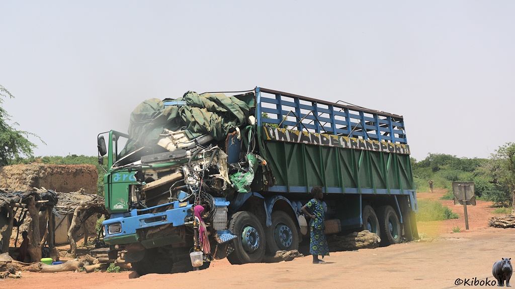 Das Bild zeigt einen grünen, vierachsigen LKW mit blauem Fahrwerk. Das Fährerhaus ist auf der Fahrerseite breitgedrückt und mit Planen zugeängt.