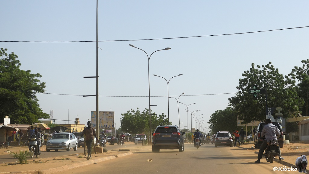 Das Bild zeigt eine staubige, vierspurige Teerstraße mit gepflasterten Mittelstreifen und Gehwegen. Windschiefe Laternen stehen auf dem Mittelstreifen. Autos, Motorräder und Fußgänger sind unterwegs.
