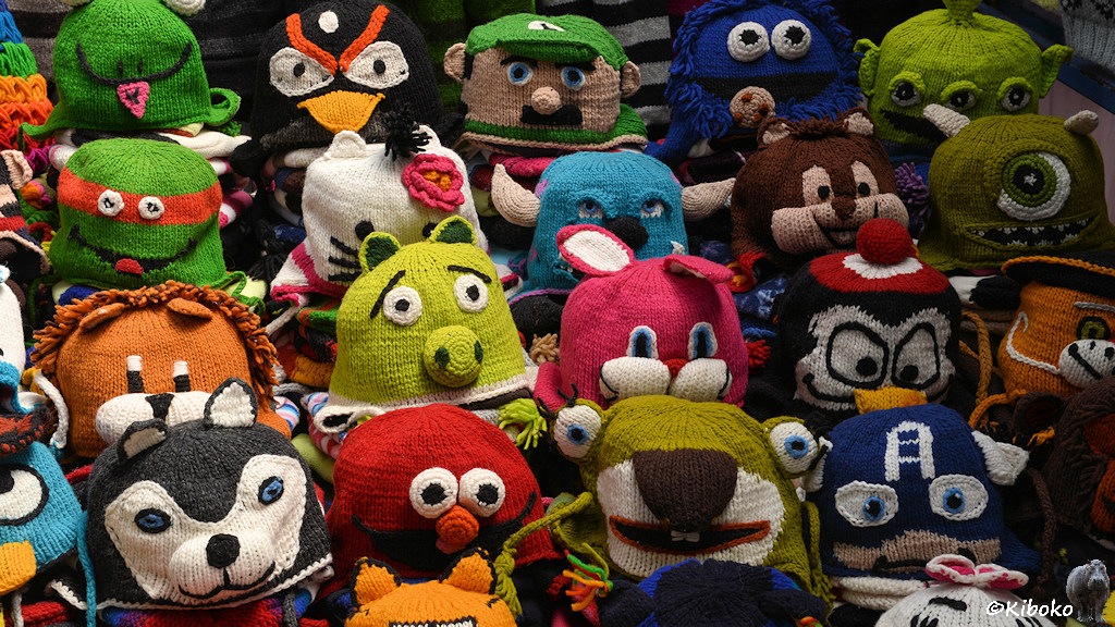Das Bild zeigt einen Stand mit gestrickten Mützen in allen Farben. Die Mützen haben Gesichter von Tieren und Außerirdischen.