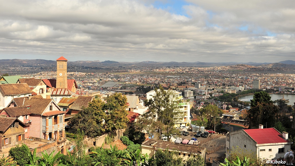 Das Bild zeigt den Blick auf eine große Stadt von einem Hügel. Am linken Bildrand stehen ein paar zweistöckige Häuser und eine Kirche mit einem viereckigen Turm aus Sandstein. Im Hintergrund breitet sich ein Häusermeer aus. Darüber ist ein wolkenverhangener Himmel.