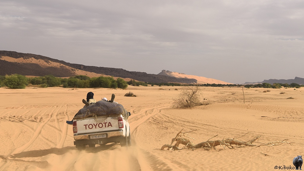 Das Bild zeigt einen vorausfahrenden weißen Geländewagen mit zwei Personen auf der vollgepackten Ladefläche. Der Wagen fährt durch losen, hellen Sand. Rechts fom Auto liegt ein vertrockneter Ast. Am linken Bildrand sind die Ausläufer einer Bergkette aus dunklen Felsen. Am Horizont ist ein Berg aus deunklen Fels, an dem sich von rechts eine hohe Düne anschmiegt.