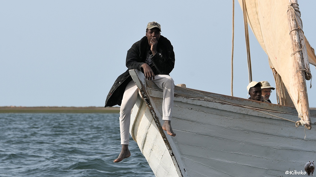 Das Bild zeigt einen Mann mit schwarzem Mantel, weißer Hose und einer olivfarbenen Baseballkappe auf der Bugspitze eines Segelbootes sitzen. Die Beine hängen über die Bordkannte.