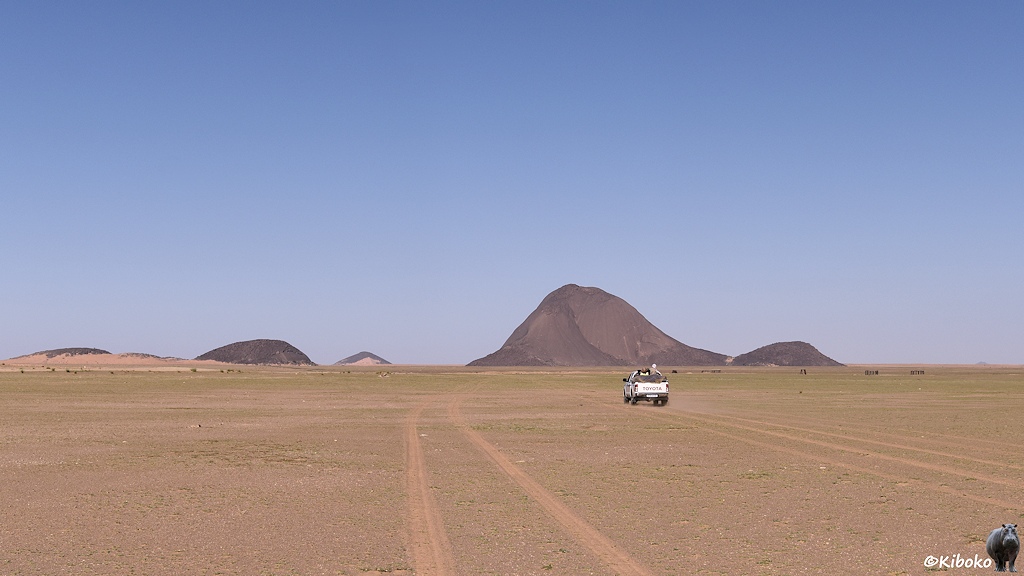 Das Bild zeigt einen weißen Geländewagen der in der Wüste auf einen rotbraunen Berg zufährt. Neben den Berg sind mehrere kleinere rotebraune Berge.