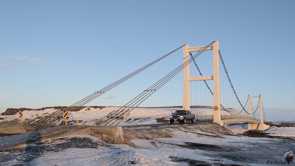 Eine Hängebrücke mit zwei hohen Pylonen aus Beton führt über einen Fluss. Ein großer Pickup hat die Brücke gerade überquert.