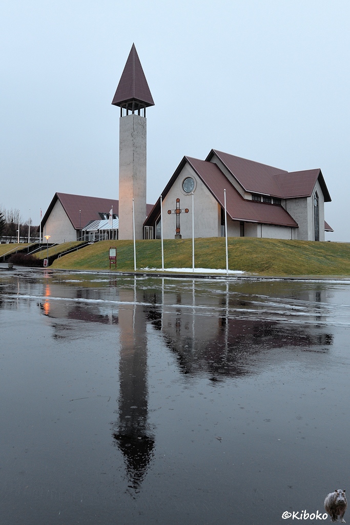 Hochformataufnahme einer modernen Kirche mit separatem, viereckigen Kirchturm aus Beton. Kirche und Turm spiegeln sich auf dem regennassen Parkplatz.
