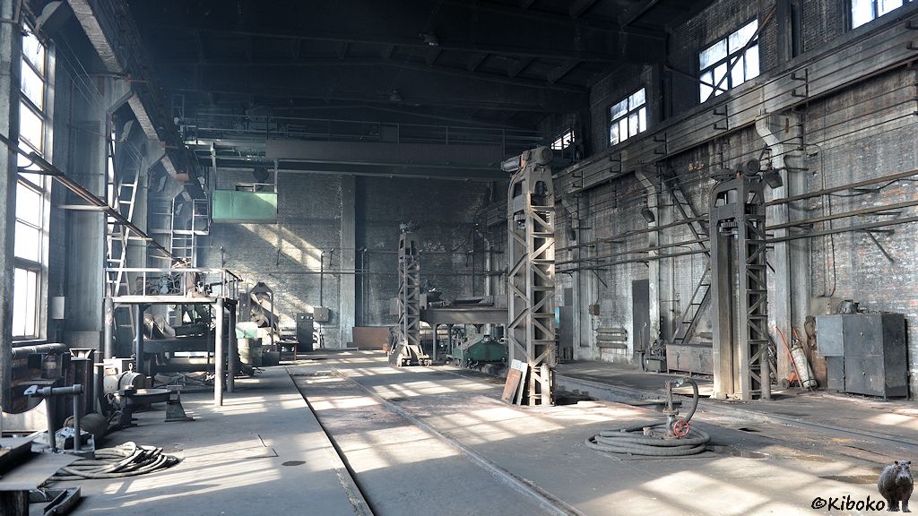 Das Bild zeigt eine leere Industriehalle mit verräucherter Decke und verstaubtem Boden. Der Deckenkran ist ebenfalls verräuchert. An der Seite stehen Maschinen und Bühnen.
