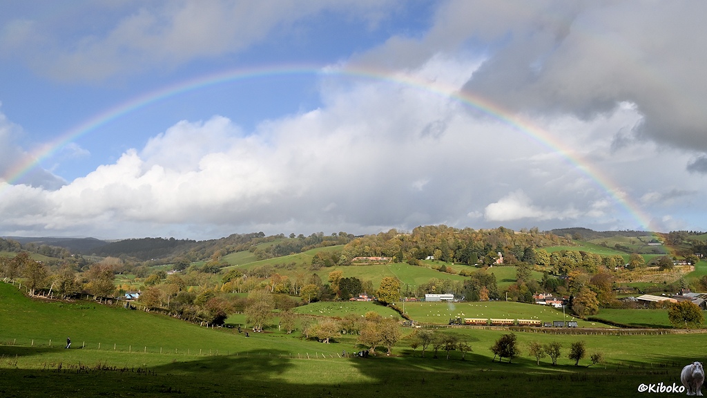 Das Bild zeigt eine Landschaftsaufnahme des selben Zuges in weiter Hügellandschaft. Ein Regenbogen überspannt Zug und Landschaft.