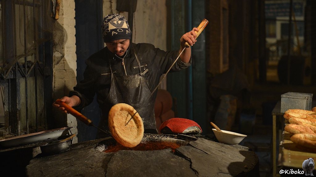 Das Bild zeigt den Bäcker mit schwarzer Jacke und schwarz-weißer Wollmütze bei der Arbeit. In den Händen hält er zwei Eisenstangen mit dem er gerade ein Fladenbrot durch die Öffnung des Steinofens gehoben hat.