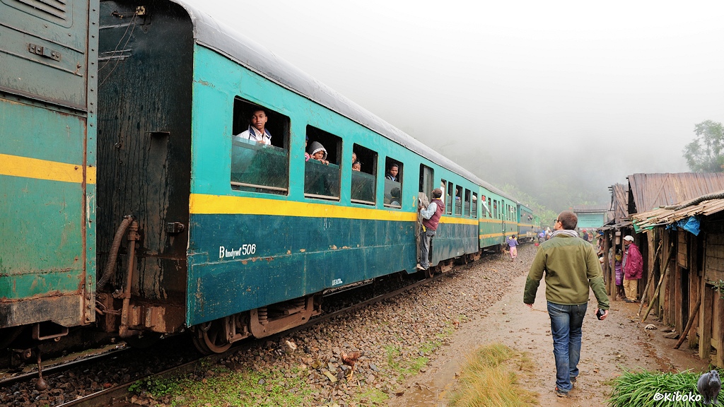 Das Bild zeigt einen dunkeltürkisen Personenwagen mit grünblauen Fensterband und gelben Trennstreifen und Mitteleinstieg. Die Fahrgäste schauen aus den offenen Fenstern. Einer steht auf dem Trittbrett.