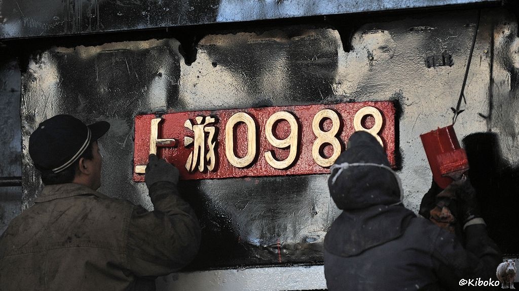Das Bild zeigt zwei Männer bei der Arbeit an einem Loknummernschild aus Messing mit der Nummer 0988. Der Linke mann renigt die Ziffern, während der rechte Mann einen großen roten Pinsel in der Hand hält.