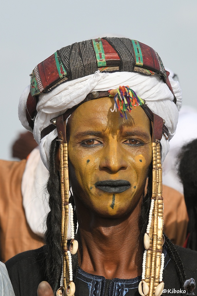 Das Bild zeigt das Porträt eines Mannes mit gelb gefäbten Gesicht, hellgelbem Streifen über Stirn, Nase und Kinn. Dazu hellgelbe Flecken mit schwarzen Punkten an den Wangen. Er trägt einen weißen Turban mit einem Schmuckgürtel. Über der Stirn ist ein Ledergürtel mit einem Büschel bunter Fransen über der Stirn. An der Seite trägt er ebenfalls goldfarbene Ketten.