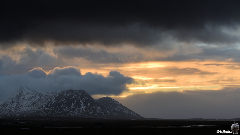 Berge ragen aus einer dunklen Eben empor. Dahinter ist eine Wolkenlücke durch die gelbes Sonnenlicht dringt. Dunkle, schwarze Wolken sind am oberen Bildrand.