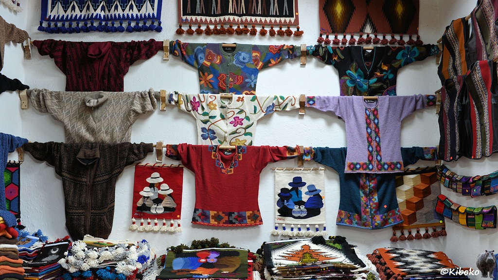 Das Bild zeigt die Rückwand des Raumes aus der Nähe. Hier hängen in drei Reihen mit jeweils drei Pullovern an der Wand. Darüber und dazwischen hängen Wanddeckchen mit Bommeln.
