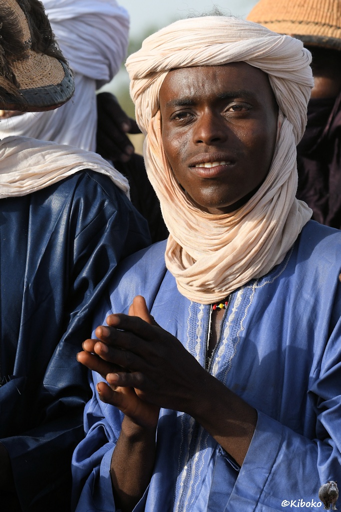 Das Bild zeigt das Porträt eines Mannes mit beigem Turban und hellblauem Gewand, der gerade in die Hände klatscht im Hochformat.