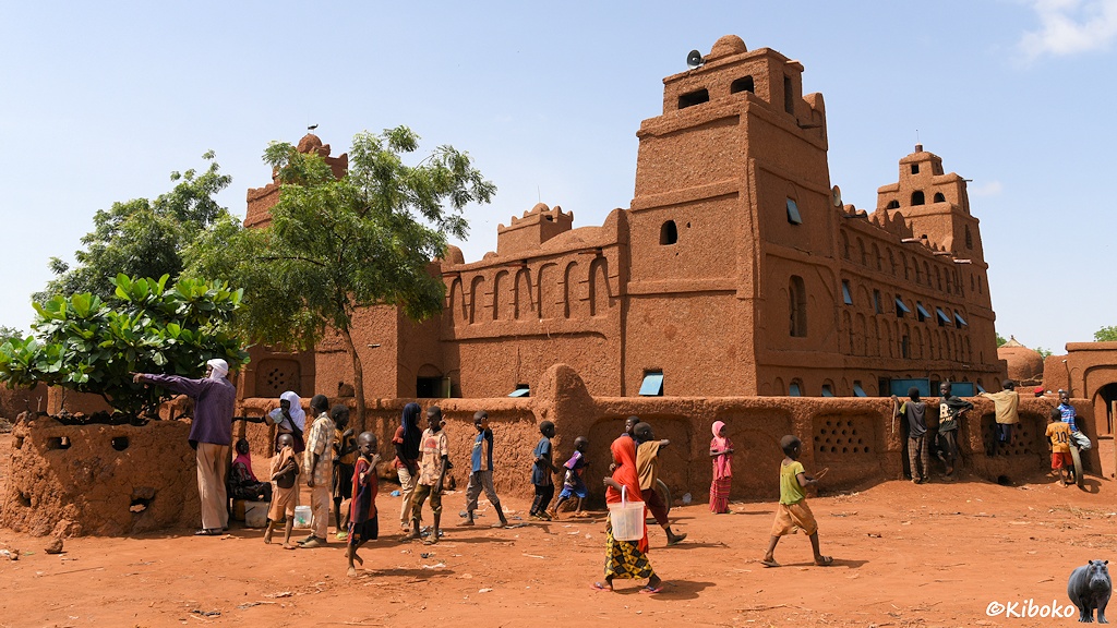 Das Bild zeigt eine viereckige Moschee mit viereckigen Ecktürmen mit der Blickrichtung von einer Ecke. Eine hohe Lehmmauer umringt die Moschee. Davor wuseln rund 20 Kinder auf einen staubigen rotbraunen Sandplatz herum.