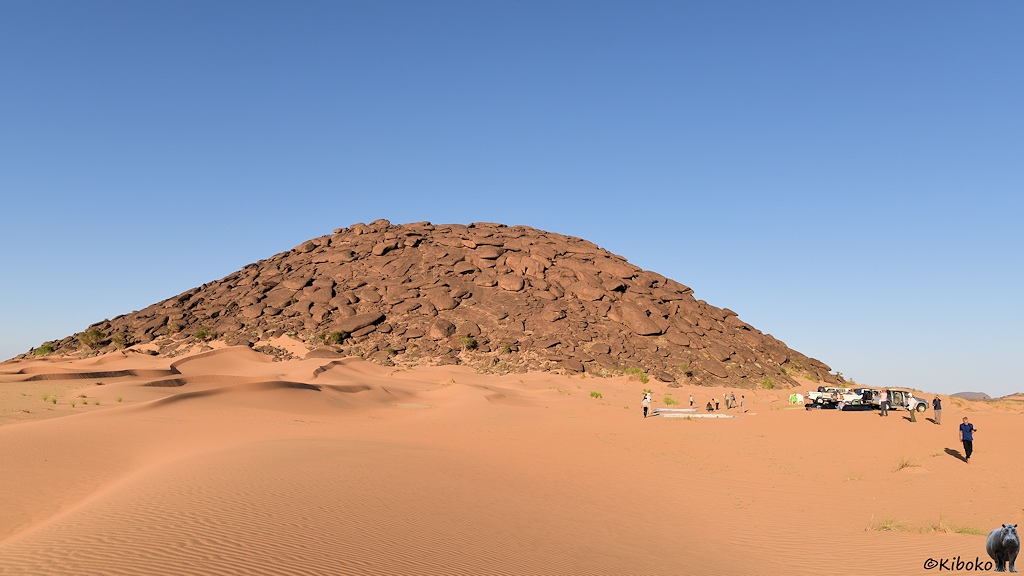 Das Bild zeigt einen rotbraunen Hügel aus großen abgerundeten Felsen. Davor sind Dünen aus blass lachsfarbenem Sand. Am rechten Bildrand stehen Geländewagen und es werden Zelte aufgebaut.