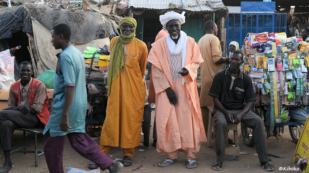 Das Bild zeigt zwei lachende ältere Männer mit Turban und orangenen und lachsfarbenen Gewandt. Sie werden von zwei sitzenden Männern an ihren Marktständen flankiert.