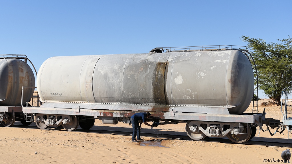 Das Bild zeigt einen hellgrauen, vierachsigen Kesselwagen. Der Kessel hat aufgeschweißte Blechflicken. Aus einem Flicken rinnt das Wasser, läuft über den Rahmen, fließst auf den Wüstenboden und bildet eine Pfütze. Ein Mann erfrischt sich am auslaufenden Wasser.