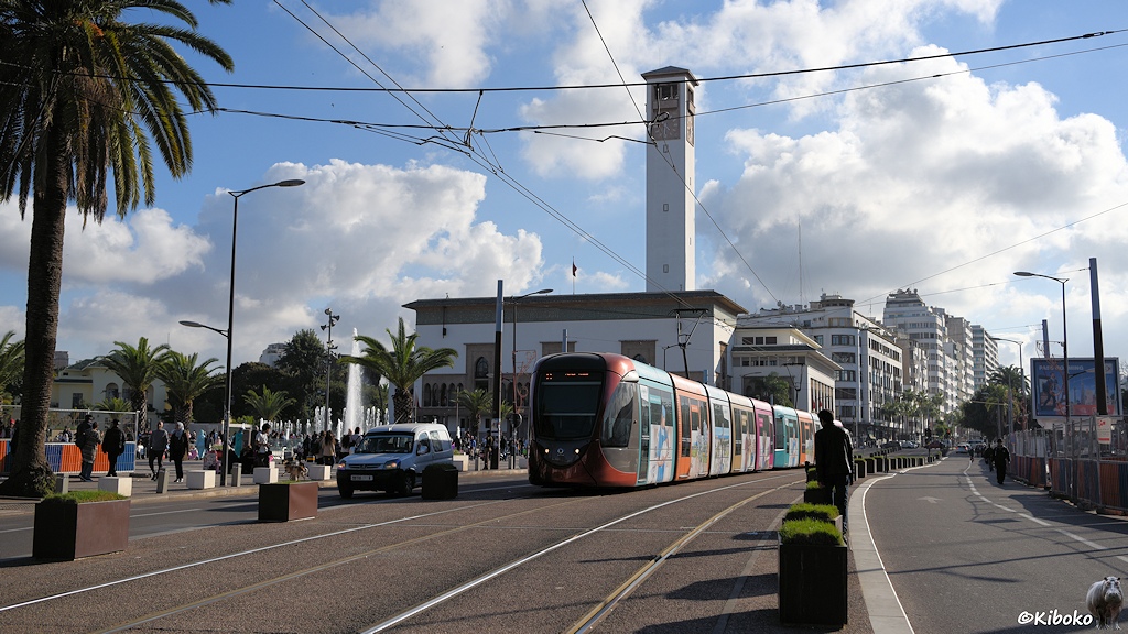 Das Bild eine moderne Straßenbahn mit rundlicher Front in Schwarz und Kupferfarben. Die Seiten sind mit bunter Reklame versehen. Die Straßenbahn fährt auf einen separaten, betonierten Fahrweg auf dem Mittelstreifen einer vierspurigen Straße. Im Hintergrund ist ein weißes Gebäude mit viereckigem Turm.