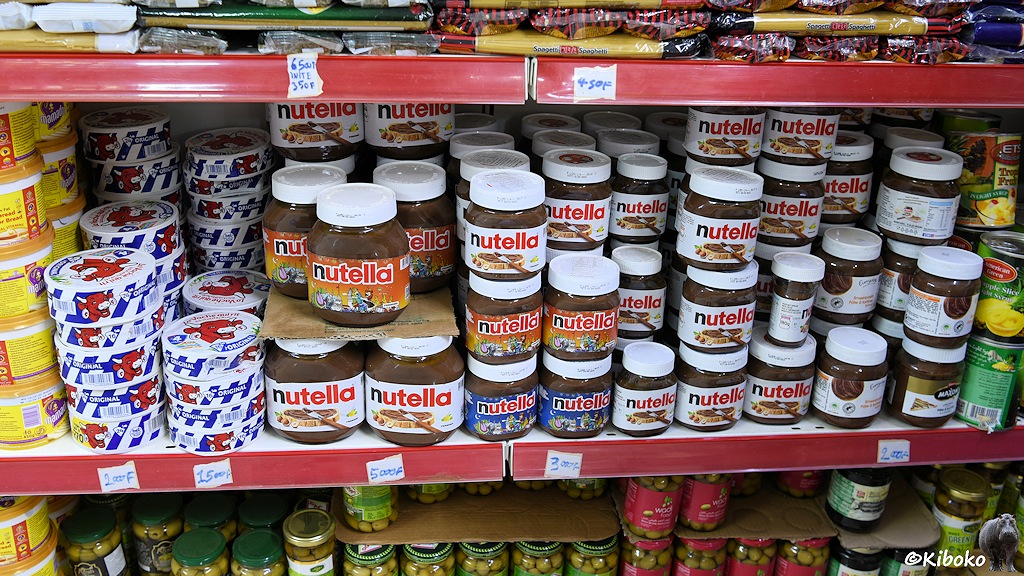 Das Bild zeigt ein Regal in einen Supermarkt. Zentral stehen ca. 100 Dosen Nutella in drei verschiedenen Größen, runde, blau-weiße Käseschachteln mit einem roten Kuhkopf, Spagetti verschiedener Hersteller.