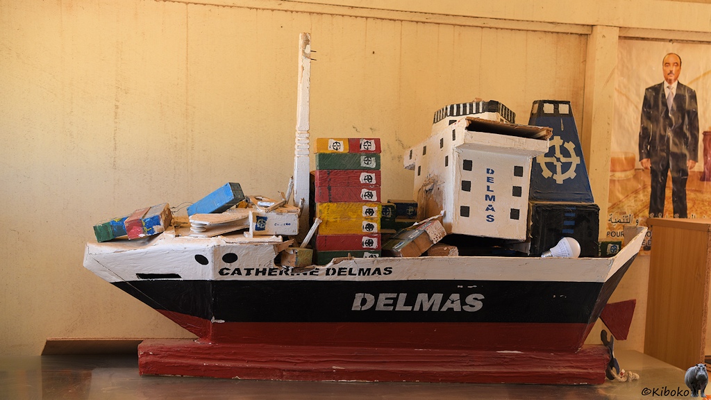 Das Bild zeigt ein Holzmodell eines weiß-schwarz-roten Containerschiffes in einem Gebäude mit beigefarbenen Wänden. Das Schiff trägt den Namen: Catherine Delmas. Viele Container liegen verstreut auf dem Deck. Das Steuerhaus mit der Brücke hat sich gelöst und steht schief auf dem Schiff. Hinter dem Schiff ist ein Plakat mit einem Mann im schwarzen Anzug.