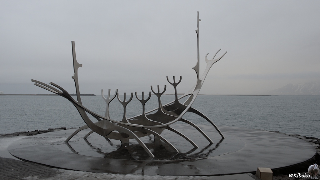 Metallstrutkur zeigt ein stilisiertes Ruderboot mit fünf Insassen. Das Denkmal steht an einem regnerrischen trüben Tag am Meer.