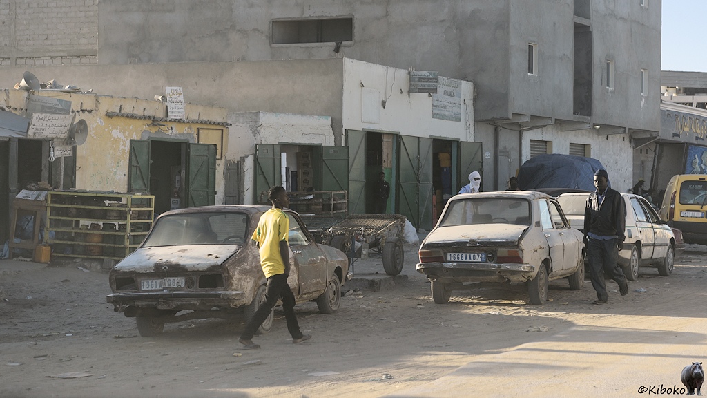 Das Bild zeigt drei PKW von hinten am staubigen Straßenrand vor einer Ladenzeile aus einstöckigen Gebäuden mit offenen grünen Türen. Beim linken braunen PKW fehlen die Rückleuchten. Der mittlere PKW ist der selbe, wie auf dem vorherigen Bild mit ausgebeulten Kofferraumdeckel. Zwei Männer laufen im Vordergrund vor den Autos.