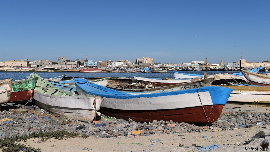 Das Bild zeigt mehrere kleine Fischerboote, die mit dem Bug am Strand liegen. Der Strand ist flächendeckend mit transparenten Plastikflaschen bedeckt. Im Hintergrund ist eine Wasserfläche und am gegenüberliegenden Ufer stehen Häuser mit Flachdach.
