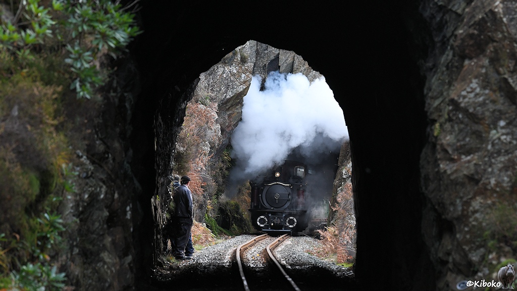 Das Bild zeigt eine Dampflok beim Verlassen eines in den Fels gehauenen Tunnels. Die Lok stößt einen weißen Dampfpilz aus. Das Bild ist durch einen weiteren Tunnel im Vordergrund fotografiert. Am Ausgang des Tunnels stehen links ein paar Personen.