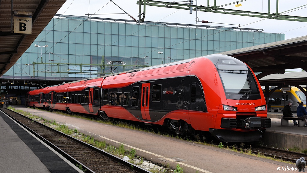 Das Bild zeigt einen modernen fünfteiligen roten Triebwagen mit einem geschwungenen, schwarz umrandeten Fensterband am Bahnsteig. Jede Triebwageneinheit hat eine rote doppeltür. Im Hintergrund ist eine Glasfront aus grünlichem Scheiben.