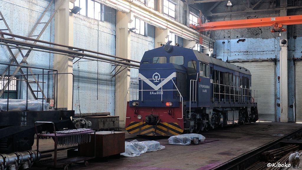 Das Bild zeigt eine dunkelblaue, sechsachsige Diesellokomotive mit zenralem Führerstand in einwer Werkhalle vor einem silbernem Rolltor. Der kurze Vorbau ist vorn. Eine rote Schlussleuchte brennt.