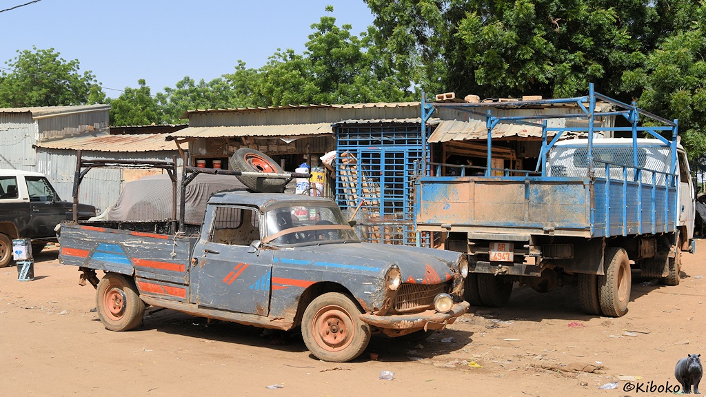 Das Bild zeigt einen dunkelblauen Peugeot-Pickup mit roten und blauen Zierstreifen auf einer staubigen Straße vor einfachen Geschäften aus Wellblech.